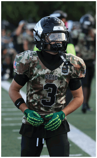 A person wearing a camouflage football uniform with the number three and a black helmet stands on a field with blurred figures in the background. Descripci n generada con IA