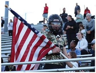 A person wearing a football helmet and camouflage jersey is holding an American flag while standing in front of bleachers with seated and standing people in the background. Descripci n generada con IA