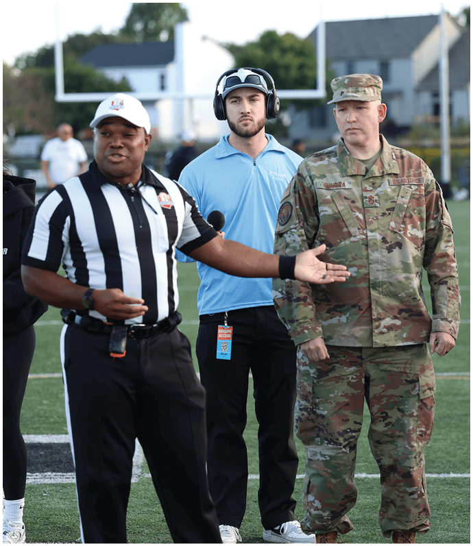 Three adults stand on a sports field, one wearing a referee uniform speaking and gesturing, another in a light blue polo with a headset, and the third in military camouflage uniform. Descripci n generada con IA