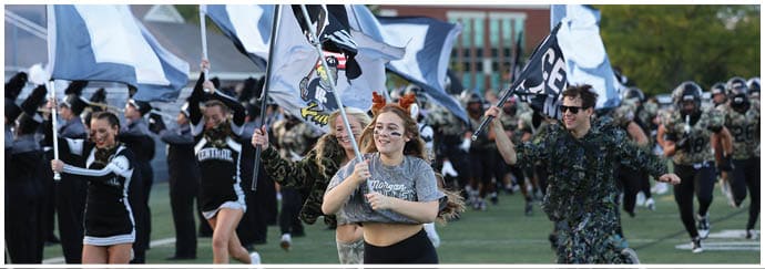 People in athletic and camouflage clothing run energetically on a sports field while holding flags, with a marching band and cheerleaders in the background. Descripci n generada con IA