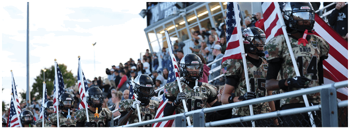 People wearing football uniforms and helmets stand behind a railing holding American flags with a crowd visible in the background. Descripci n generada con IA