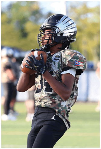 A person wearing a camouflage football jersey and black pants is holding a football while standing on a grassy field during a game. Descripci n generada con IA