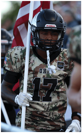 A person wearing a camouflage football uniform with the number 17 and a black helmet is holding an American flag. Descripci n generada con IA