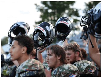 People in camouflage uniforms are holding up black football helmets with faceguards in an outdoor setting. Descripci n generada con IA