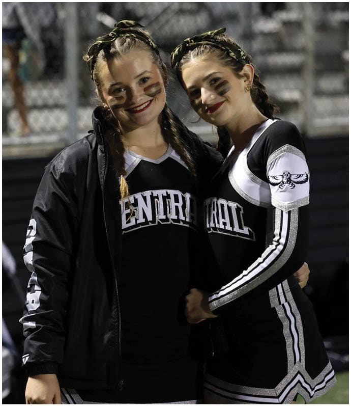 Two people wearing cheerleading uniforms with braided hair and face paint stand closely together smiling on a sports field at night. Descripci n generada con IA