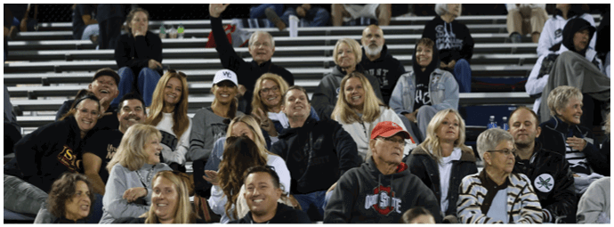 A group of people sitting closely together on bleachers at an outdoor event, some smiling and engaged in conversation, with casual clothing and a few wearing hats. Descripci n generada con IA