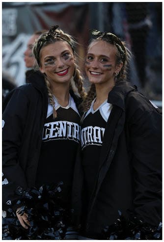 Two adults wearing matching cheerleading uniforms and jackets smile with braided hair and face paint while standing close together outdoors. Descripci n generada con IA