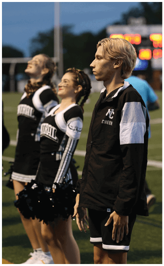 Three people in cheerleading uniforms stand on a sports field with a scoreboard visible in the background during an evening event. Descripci n generada con IA