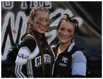 Two people wearing cheerleading uniforms with face paint smile and pose together in front of a blurred background. Descripci n generada con IA