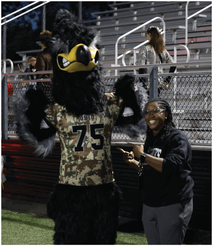 A person in a black bird mascot costume wearing a camouflage sports jersey with the number 75 stands on a football field near bleachers with people seated and standing. Descripci n generada con IA