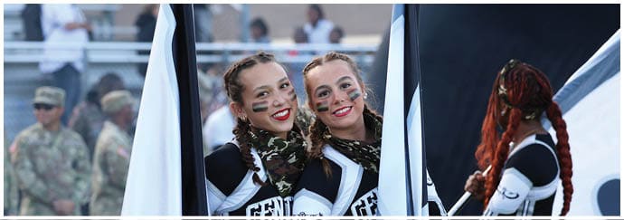 Two adults wearing cheerleading uniforms and face paint smile while holding large flags on a sports field with people in military uniforms in the background. Descripci n generada con IA