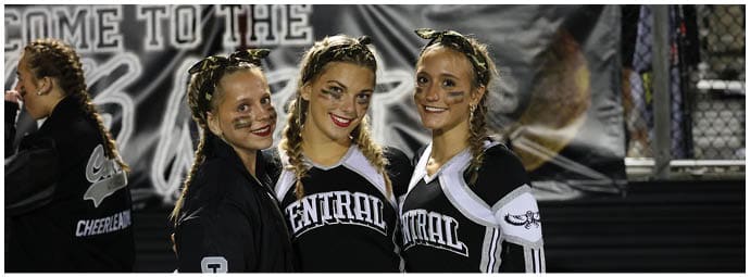 Three people wearing matching cheerleading uniforms with “Central" on the front and face paint are standing close together and smiling at an outdoor event. Descripci n generada con IA