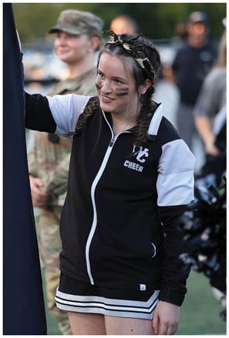 A person wearing a cheerleading outfit with face paint and braided hair is holding a large dark flag while another person in military uniform stands blurred in the background. Descripci n generada con IA