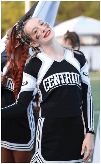 A person in a black and white cheerleading uniform holding a large flag smiles while standing on a field with other similarly dressed people holding flags in the background. Descripci n generada con IA