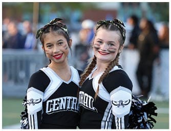 Two people in matching cheerleading uniforms with braided hair and face paint smile while standing close together on a sports field. Descripci n generada con IA