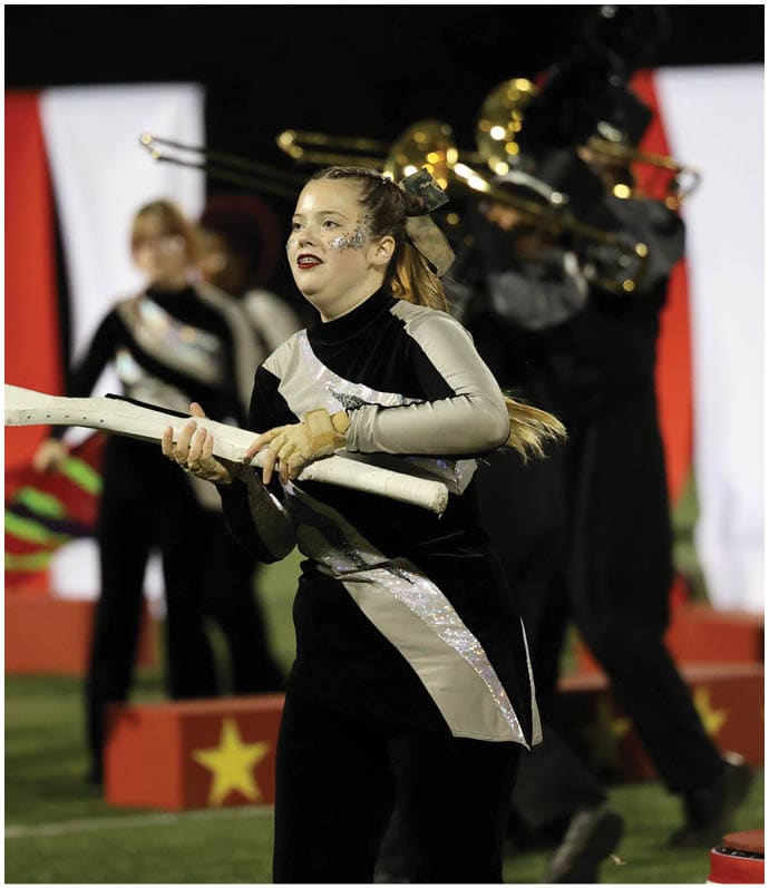 A person in a black and silver uniform is performing with a white rifle prop on a field while other band members play brass instruments in the background. Descripci n generada con IA