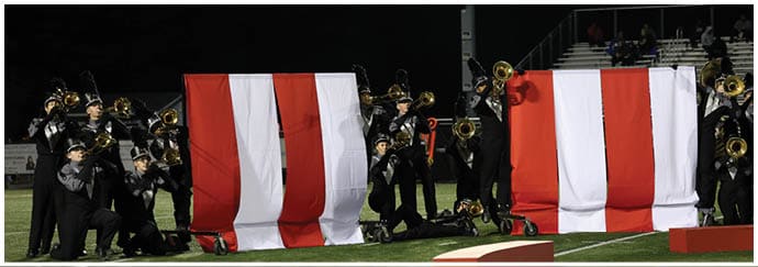People in marching band uniforms are playing brass instruments behind two large red and white striped banners on a field at night. Descripci n generada con IA