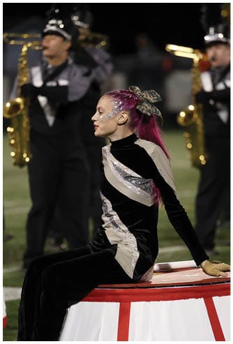 A person with bright pink hair tied with a camouflage bow and glitter on their face is wearing a black and silver costume and appears to be performing on a field with a blurred musician in the background. Descripci n generada con IA