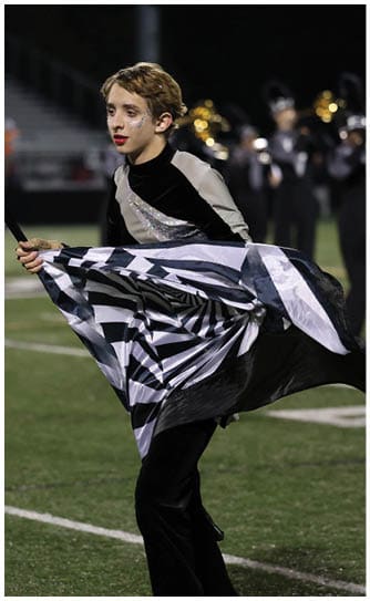 A person in a black and silver costume is holding and waving a black and white striped flag on a sports field during a nighttime event. Descripci n generada con IA