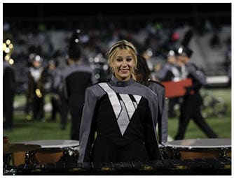 A person wearing a gray and black marching band uniform with a silver diagonal stripe is smiling while standing on a field with other similarly dressed people in the background. Descripci n generada con IA