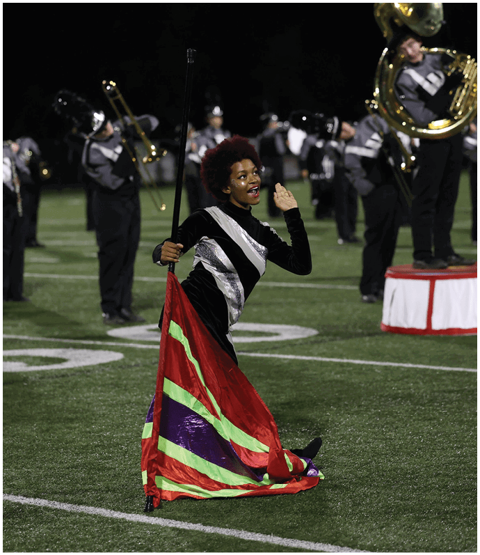 A person with short curly hair wearing a black and silver uniform is kneeling on a football field holding a colorful flag while a marching band plays brass instruments in the background. Descripci n generada con IA
