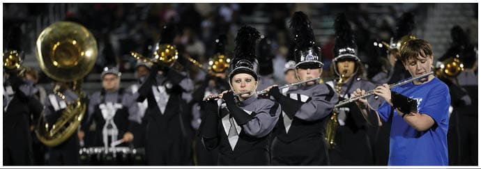 People in a marching band uniform play flutes and brass instruments on a field at night. Descripci n generada con IA