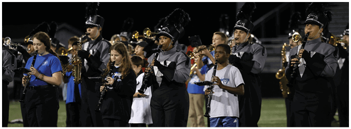 People in a marching band uniform and casual clothing are standing in a row playing clarinets on a field at night. Descripci n generada con IA