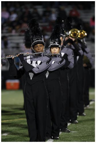 People in marching band uniforms with tall feathered hats are playing flutes in formation on a field. Descripci n generada con IA