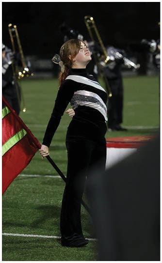 A person in a black and silver uniform stands on a field holding a red and green flag with a marching band playing brass instruments in the background. Descripci n generada con IA