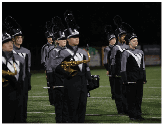 People in marching band uniforms stand in formation on a field at night holding musical instruments. Descripci n generada con IA