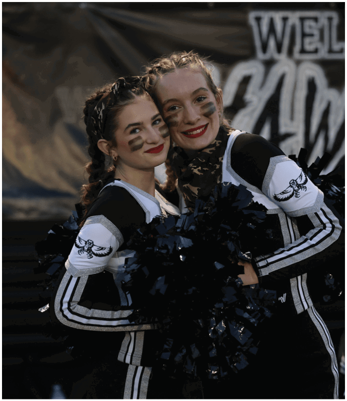 Two people in matching cheerleading uniforms with face paint smile and pose closely together holding black pom-poms. Descripci n generada con IA