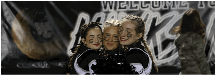 Three people in cheerleading uniforms with face paint are smiling and hugging each other on a sports field. Descripci n generada con IA