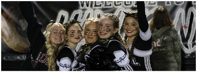 Five people wearing cheerleading uniforms and face paint smile and pose with their arms raised in front of a welcome banner at an outdoor event. Descripci n generada con IA