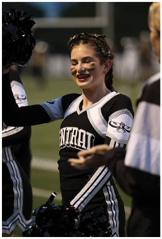People in cheerleading uniforms are holding pom-poms and interacting on a sports field during an event. Descripci n generada con IA