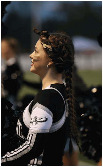 A person with long braided hair wearing a camouflage headband and a black and white cheerleading uniform is smiling and looking to the side outdoors. Descripci n generada con IA