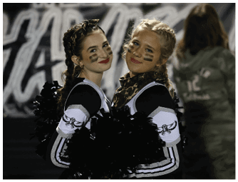 Two adults dressed in matching cheerleading uniforms with face paint and braided hair smile while holding black pom-poms at an outdoor event. Descripci n generada con IA