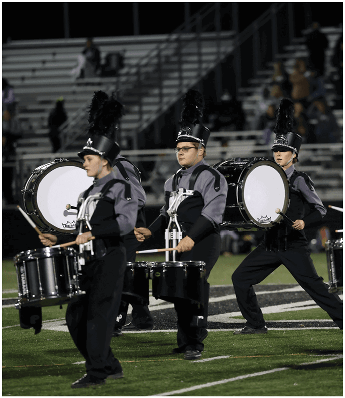 People in marching band uniforms with tall feathered hats are playing drums on a football field with empty bleachers in the background. Descripci n generada con IA