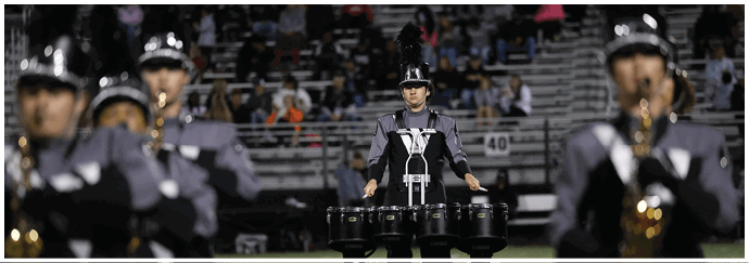 Three people in marching band uniforms with hats and plumes are playing instruments in front of a crowd seated on bleachers at night. Descripci n generada con IA