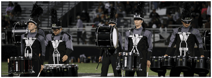 Five people wearing matching band uniforms and tall feathered hats are playing bass drums in a stadium with bleachers and a crowd in the background. Descripci n generada con IA