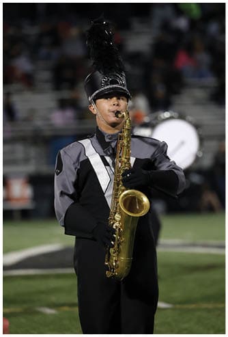 A person in a marching band uniform with a tall feathered hat is playing a saxophone on a field. Descripci n generada con IA