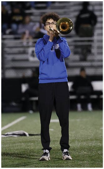A person wearing a blue sweatshirt and black pants is playing a trumpet on a sports field with other trumpet players lined up nearby and spectators seated in the background. Descripci n generada con IA