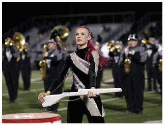 A person with long pink hair in a black and silver uniform is holding a white rifle prop on a field with a marching band playing instruments in the background. Descripci n generada con IA