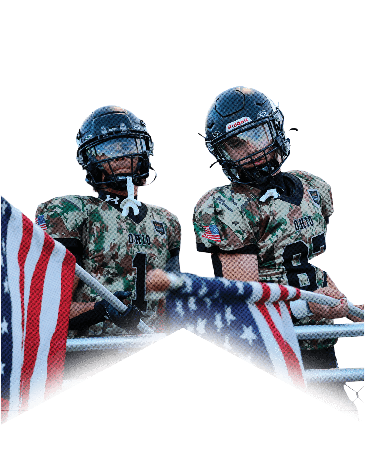 Two people in camouflage football uniforms and helmets stand behind a railing holding American flags with bleachers and a crowd in the background. Descripci n generada con IA
