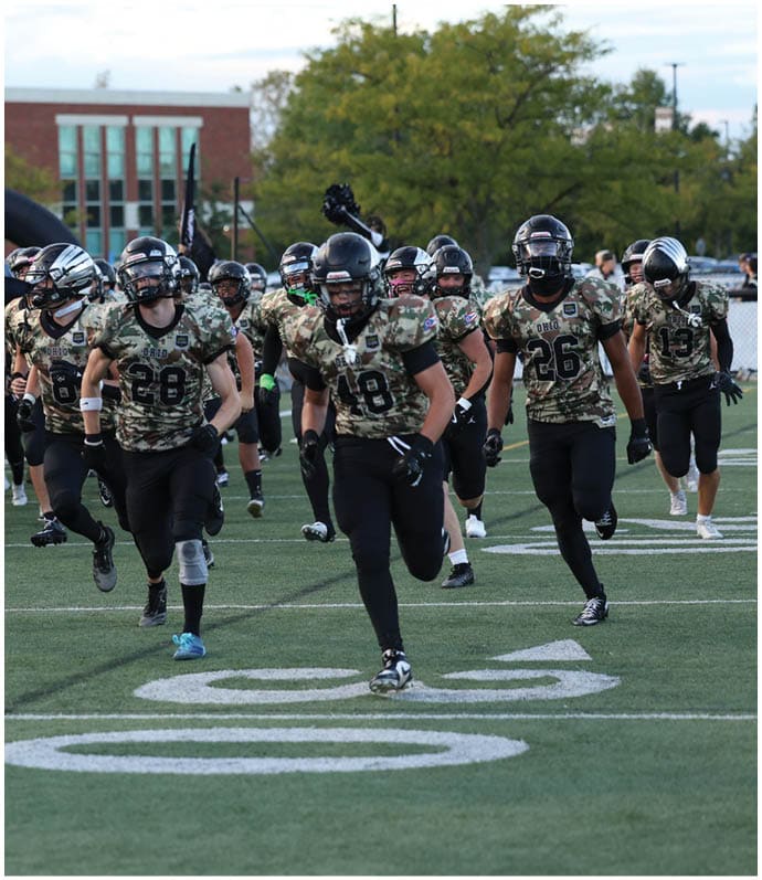 A group of people in camouflage sports uniforms and helmets are running onto a football field with one person holding a large flag and a cheerleader running alongside them. Descripci n generada con IA