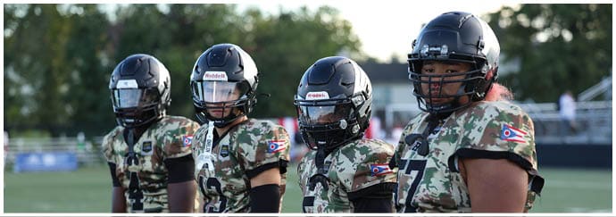 Four people wearing camouflage football uniforms and helmets stand in a row on a sports field. Descripci n generada con IA