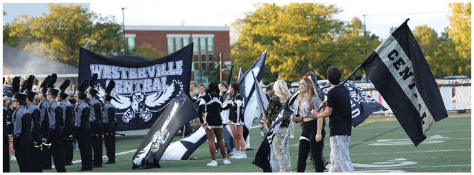 People in marching band uniforms and cheerleading outfits stand on a football field holding large black and white flags with “Westerville Central" written on them during an outdoor event. Descripci n generada con IA