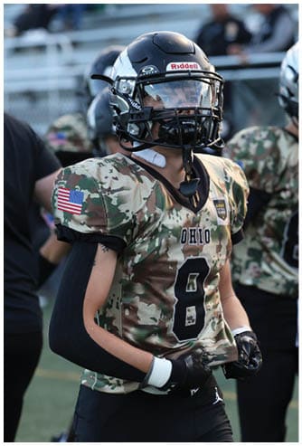 A person wearing a camouflage football uniform with the word Ohio and the number 8 on the front stands on a field with blurred spectators in the background. Descripci n generada con IA