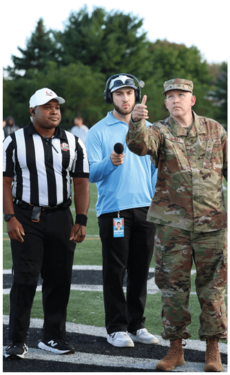 A person in military uniform is giving a thumbs-up gesture while standing on a sports field next to a person holding a microphone and wearing a headset and a blue shirt. Descripci n generada con IA
