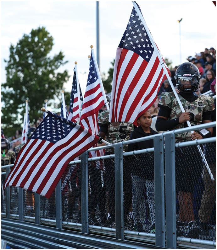People in camouflage uniforms holding American flags stand behind a metal fence with a crowd in the background. Descripci n generada con IA