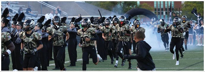A group of people wearing football uniforms and helmets are running onto a field through smoke with a crowd and cheerleaders in the background. Descripci n generada con IA
